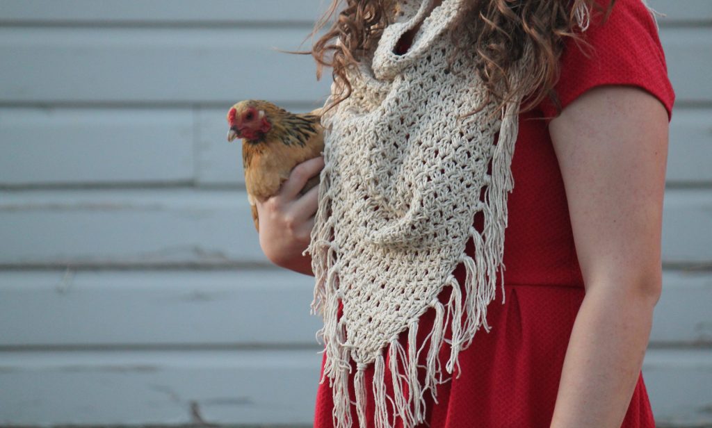 Model in red dress and fringed crochet cowl holding a chicken.